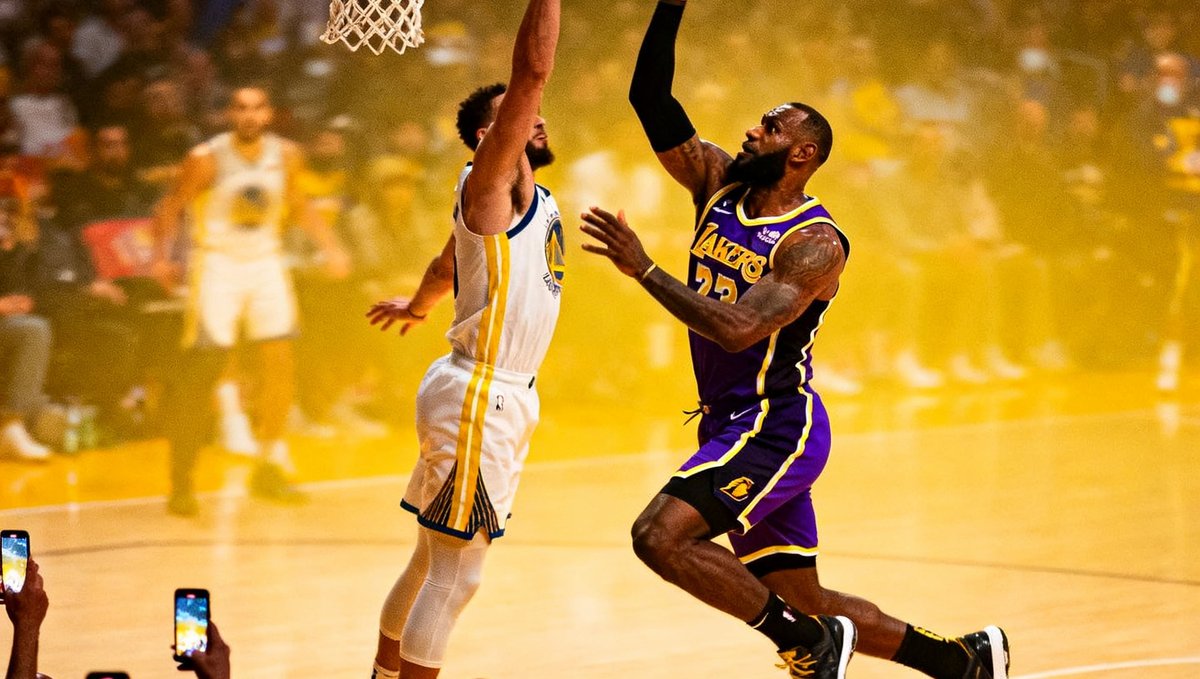 NBA赛场上湖人队球员飞速快攻，腾空而起准备单手劈扣，防守球员奋力起跳拦截，篮筐后方观众纷纷举起手机拍摄